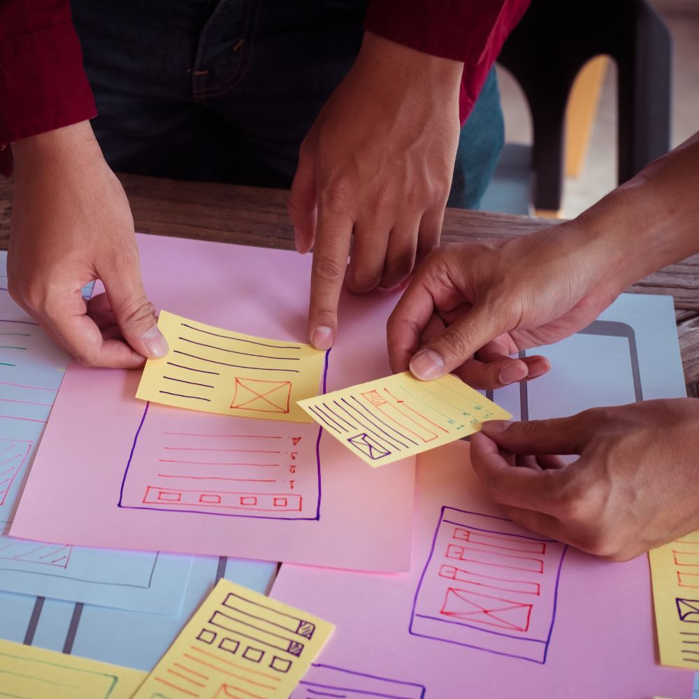 preparation-exposition-guide Trois mains collaborant sur des notes colorées et des plans, symbolisant la préparation méthodique pour une exposition réussie.